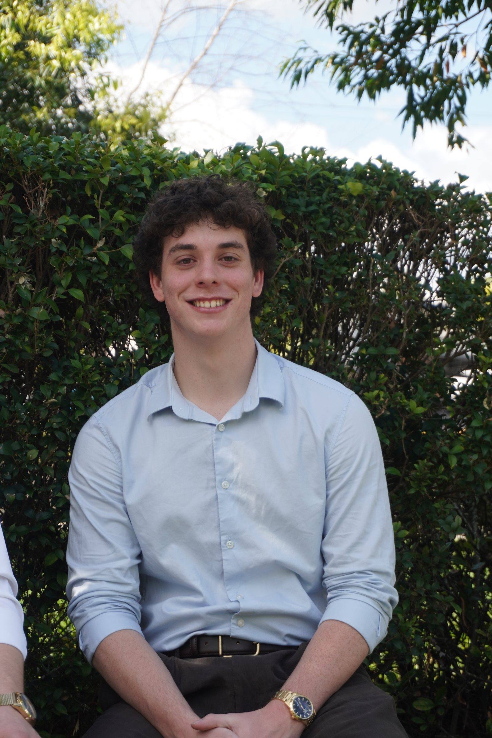 Levi, with curly dark hair and a light-blue shirt, seated in front of a green hedge